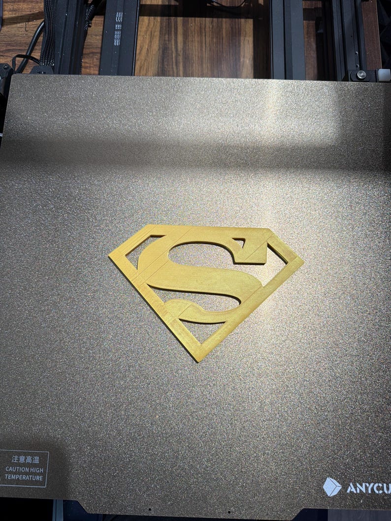 May include: A gold-colored 3D printed Superman logo emblem centered on a dark gray textured build plate. The build plate features a white warning label with the text CAUTION HIGH TEMPERATURE and the brand name ANYCU visible in the corner.