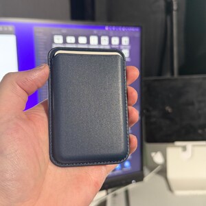 May include: A navy blue leather card holder with a stitched border. The card holder has a rectangular shape with rounded corners and a cream-colored interior. The product is held in a person's hand.