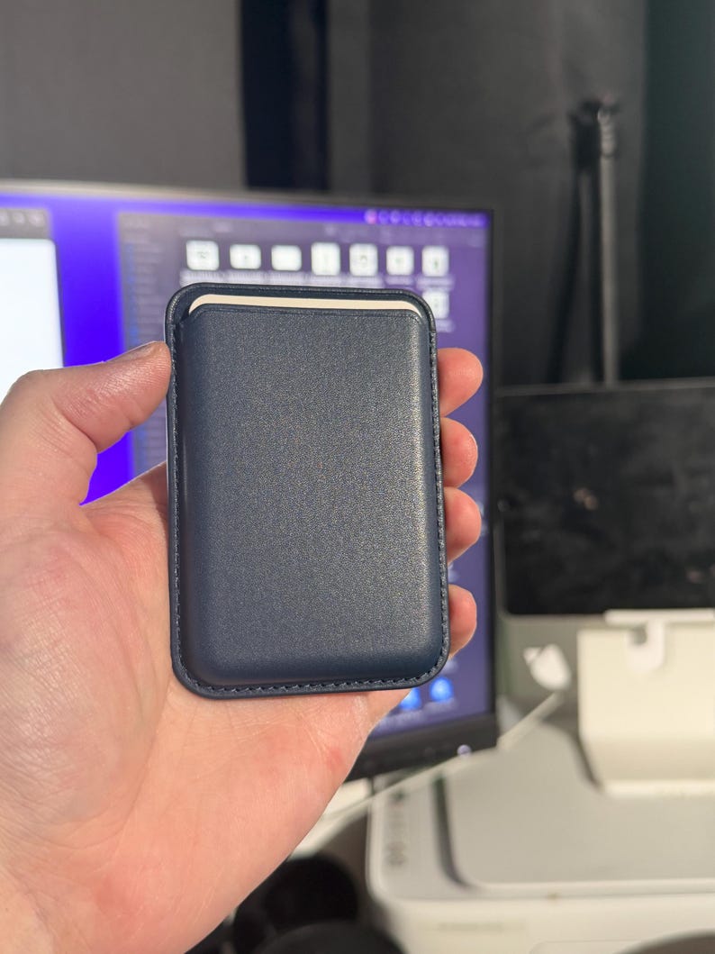 May include: A navy blue leather card holder with a stitched border. The card holder has a rectangular shape with rounded corners and a cream-colored interior. The product is held in a person's hand.