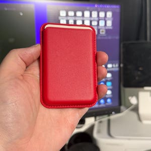 May include: A red leather phone case with a white interior. The case is rectangular with rounded corners and a stitched border. The case is held in a hand.