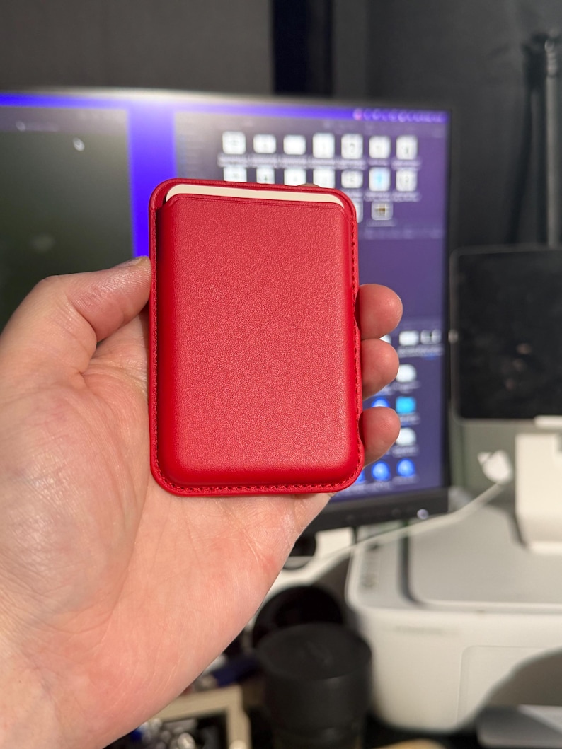 May include: A red leather phone case with a white interior. The case is rectangular with rounded corners and a stitched border. The case is held in a hand.