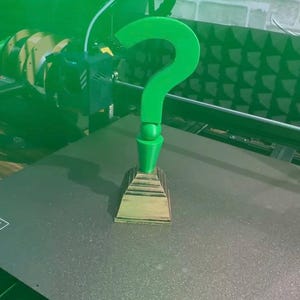 May include: A 3D-printed green question mark, a symbol of the Riddler, stands on a tiered base. The object is being created by a 3D printer. The text "RICHIE'S LENS 3D" is visible in the background.