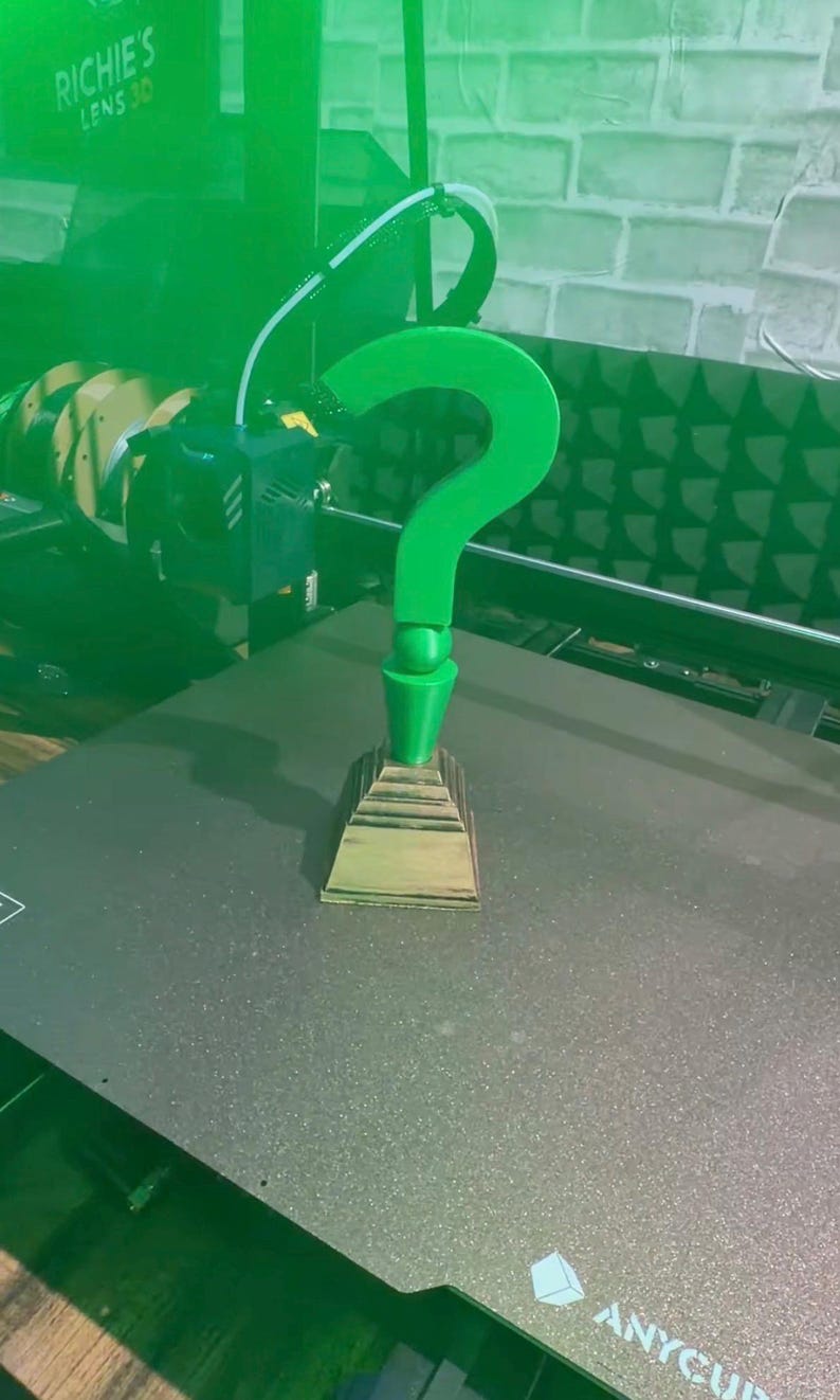 May include: A 3D-printed green question mark, a symbol of the Riddler, stands on a tiered base. The object is being created by a 3D printer. The text "RICHIE'S LENS 3D" is visible in the background.