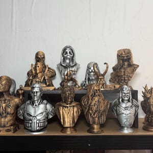 May include: A collection of character busts, including Spider-Man, The Punisher, and others. The busts are in various metallic colors, such as gold and silver, and are displayed on a shelf. The collection features detailed designs and textures.