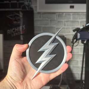 May include: A silver and black Flash logo emblem. The emblem features a silver lightning bolt design set against a black circular background, with a silver border. The emblem is held in a hand.