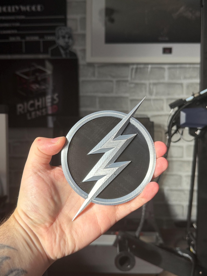 May include: A silver and black Flash logo emblem. The emblem features a silver lightning bolt design set against a black circular background, with a silver border. The emblem is held in a hand.