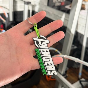 K&ouml;nnte beinhalten: Ein schwarz-wei&szlig;er Avengers-Schl&uuml;sselanh&auml;nger mit dem Wort "DOOMSDAY" in Gr&uuml;n. Der Schl&uuml;sselanh&auml;nger ist an einer gr&uuml;nen Schlaufe und einem silbernen Schl&uuml;sselring befestigt. Das Avengers-Logo befindet sich oben auf dem Schl&uuml;sselanh&auml;nger.