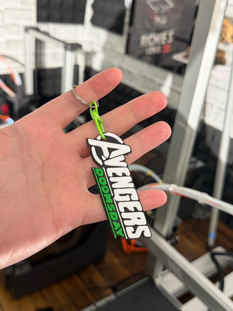 K&ouml;nnte beinhalten: Ein schwarz-wei&szlig;er Avengers-Schl&uuml;sselanh&auml;nger mit dem Wort "DOOMSDAY" in Gr&uuml;n. Der Schl&uuml;sselanh&auml;nger ist an einer gr&uuml;nen Schlaufe und einem silbernen Schl&uuml;sselring befestigt. Das Avengers-Logo befindet sich oben auf dem Schl&uuml;sselanh&auml;nger.