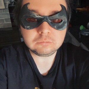 May include: A black eye mask with pointed upper edges and textured, reddish-brown lenses covering the eye area. The mask features a contoured shape that fits across the bridge of the nose and around the eyes, creating a distinct superhero-inspired look.