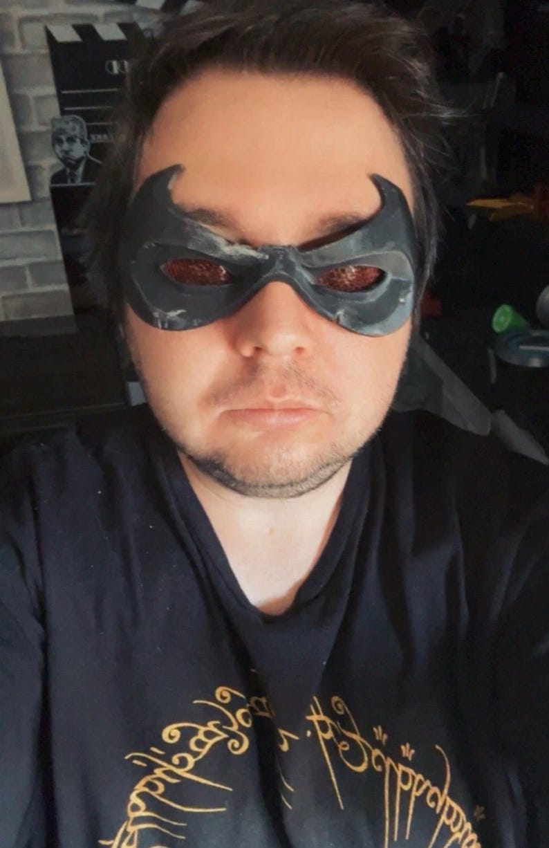 May include: A black eye mask with pointed upper edges and textured, reddish-brown lenses covering the eye area. The mask features a contoured shape that fits across the bridge of the nose and around the eyes, creating a distinct superhero-inspired look.