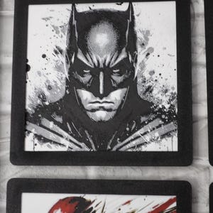 May include: A square black-framed wall art piece featuring a black and white ink-style illustration of Batman. The character is depicted from the chest up with a focused expression, surrounded by abstract paint splatters and textured brush strokes.