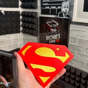 May include: A diamond-shaped emblem featuring the iconic red and yellow Superman logo. The design consists of a bold red outer border and a central red S-shape, set against a textured yellow background, creating a distinct layered appearance.