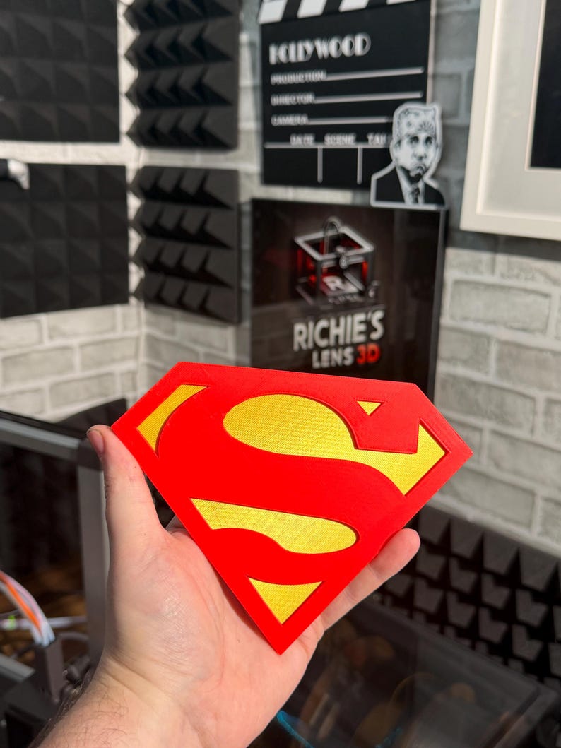 May include: A diamond-shaped emblem featuring the iconic red and yellow Superman logo. The design consists of a bold red outer border and a central red S-shape, set against a textured yellow background, creating a distinct layered appearance.