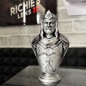 May include: A silver-colored bust of a king wearing a crown and armor. The bust is 3D printed and sits on a round base. The background includes a sign that reads "RICHIE'S LENS 3D".