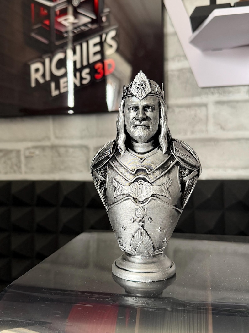 May include: A silver-colored bust of a king wearing a crown and armor. The bust is 3D printed and sits on a round base. The background includes a sign that reads "RICHIE'S LENS 3D".