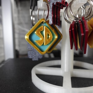 May include: A display of keychains featuring a gold and teal diamond-shaped charm with the letters "SD" in gold. Other keychains include red, black, and silver designs. The keychains are hanging on a white display stand.