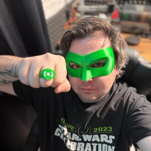 Puede incluir: Una persona con una m&aacute;scara verde y un anillo verde. La persona lleva una camiseta negra con el texto "ELOP 2023 STAR WARS CELEBRATION". El fondo est&aacute; borroso, con enfoque en la persona y los accesorios.