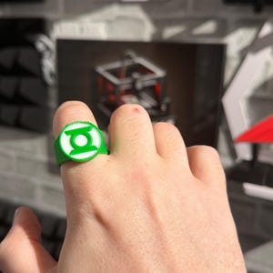 Puede incluir: Un anillo verde y blanco con el logotipo de Green Lantern. El anillo es circular con un emblema en relieve en el centro. El anillo se lleva en un dedo.