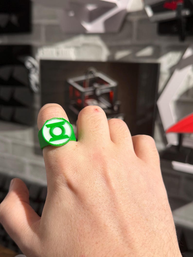 Puede incluir: Un anillo verde y blanco con el logotipo de Green Lantern. El anillo es circular con un emblema en relieve en el centro. El anillo se lleva en un dedo.