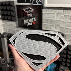 May include: A shield-shaped emblem featuring the letter S in a stylized font. The emblem has a light gray textured surface with a repeating pattern and deep black recessed sections that create a high-contrast layered appearance.