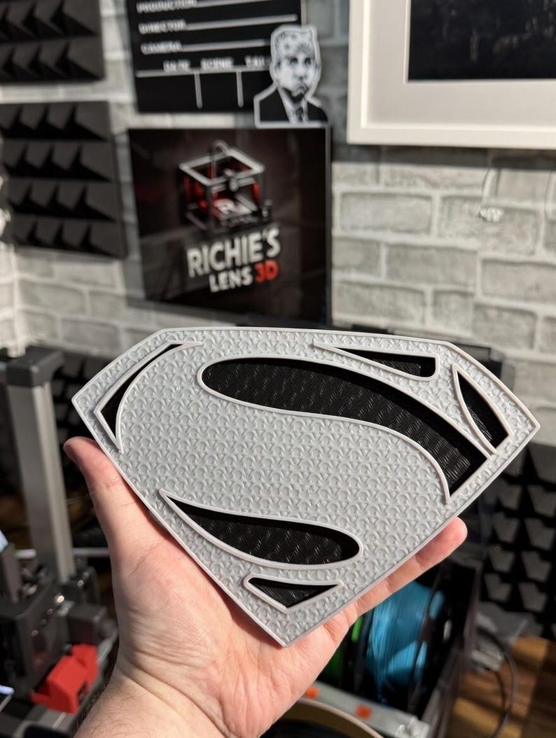 May include: A shield-shaped emblem featuring the letter S in a stylized font. The emblem has a light gray textured surface with a repeating pattern and deep black recessed sections that create a high-contrast layered appearance.