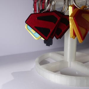 K&ouml;nnte beinhalten: Eine Pr&auml;sentation von Superman-Logo-Schl&uuml;sselanh&auml;ngern in Rot, Schwarz und Gold. Die Schl&uuml;sselanh&auml;nger sind an silbernen Ringen befestigt und h&auml;ngen an einem wei&szlig;en, mehrst&ouml;ckigen Pr&auml;sentationsst&auml;nder. Das Superman-Logo ist rautenf&ouml;rmig.