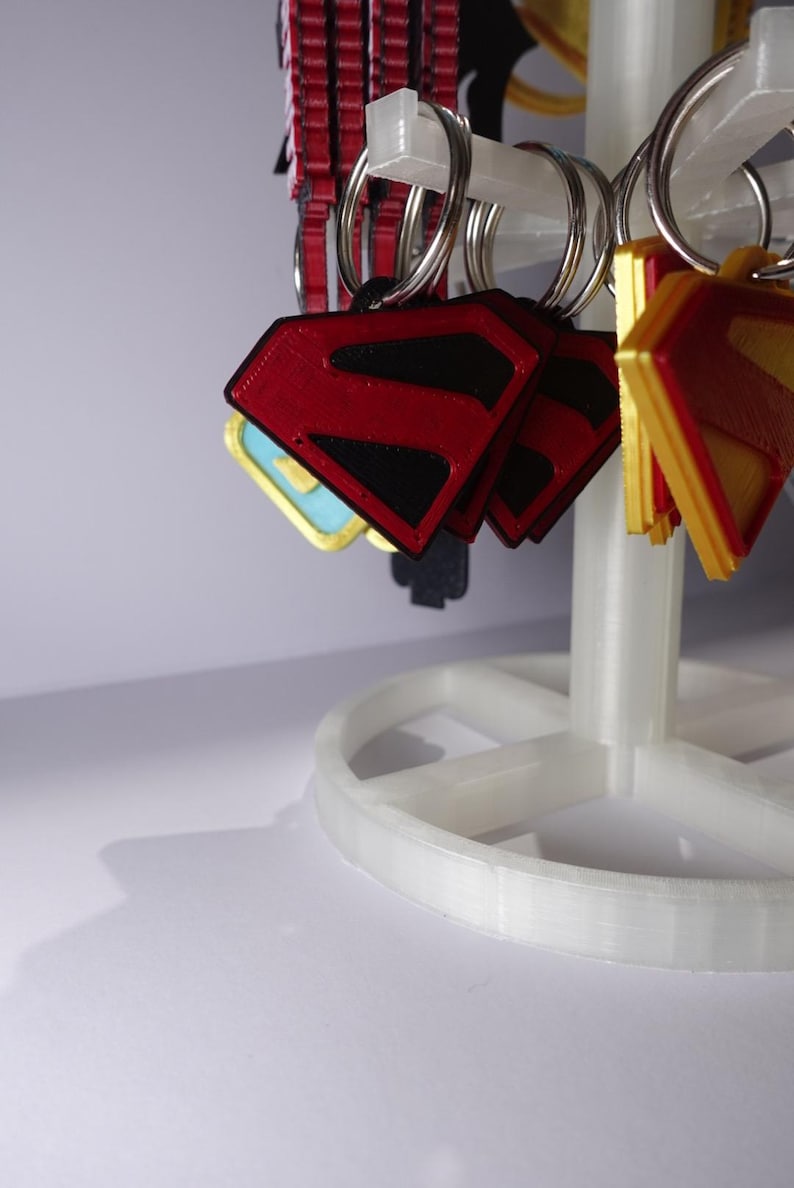 K&ouml;nnte beinhalten: Eine Pr&auml;sentation von Superman-Logo-Schl&uuml;sselanh&auml;ngern in Rot, Schwarz und Gold. Die Schl&uuml;sselanh&auml;nger sind an silbernen Ringen befestigt und h&auml;ngen an einem wei&szlig;en, mehrst&ouml;ckigen Pr&auml;sentationsst&auml;nder. Das Superman-Logo ist rautenf&ouml;rmig.