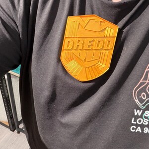 May include: A gold-colored Dredd badge is pinned to a black t-shirt. The badge is shield-shaped with the word "DREDD" in bold letters. The shirt has a graphic on the lower right side. The badge is a fan-made item.