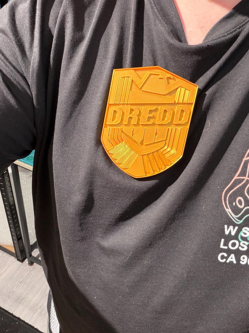 May include: A gold-colored Dredd badge is pinned to a black t-shirt. The badge is shield-shaped with the word "DREDD" in bold letters. The shirt has a graphic on the lower right side. The badge is a fan-made item.