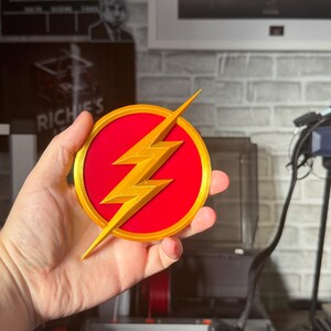 May include: A round, red and gold emblem featuring a gold lightning bolt. The emblem is a 3D-printed design, likely a fan-made item or collectible. The background includes a brick wall and a framed picture.