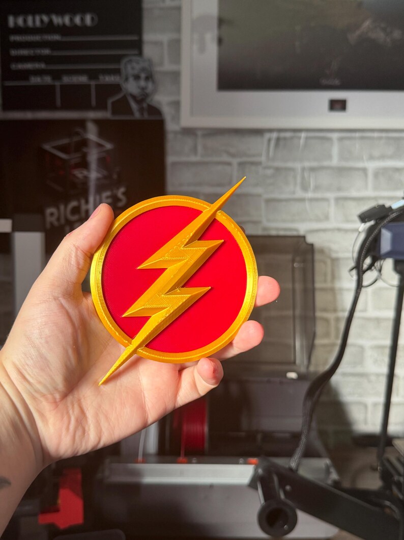 May include: A round, red and gold emblem featuring a gold lightning bolt. The emblem is a 3D-printed design, likely a fan-made item or collectible. The background includes a brick wall and a framed picture.