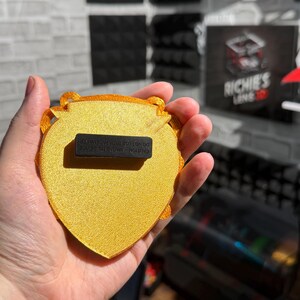 May include: A gold-colored, heart-shaped magnet with a textured surface. The magnet has a decorative border and a black rectangular magnet attached to the back. The text "CAUTION - MAGNETIC DEVICE DO NOT USE WITH PACEMAKER" is printed on the back.