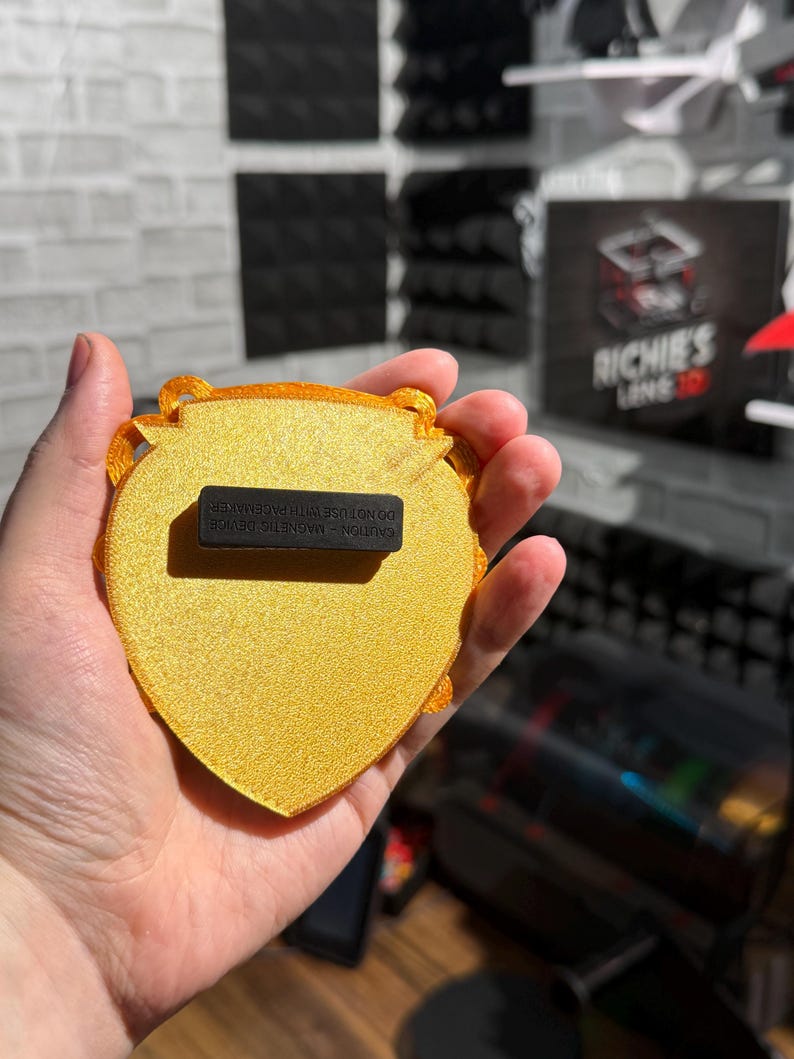 May include: A gold-colored, heart-shaped magnet with a textured surface. The magnet has a decorative border and a black rectangular magnet attached to the back. The text "CAUTION - MAGNETIC DEVICE DO NOT USE WITH PACEMAKER" is printed on the back.