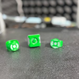 Puede incluir: Tres objetos impresos en 3D con el tema de Green Lantern, verdes y blancos. Los objetos incluyen dos anillos y un cubo, todos con el logotipo de Green Lantern. Los art&iacute;culos se muestran sobre una superficie gris texturizada.