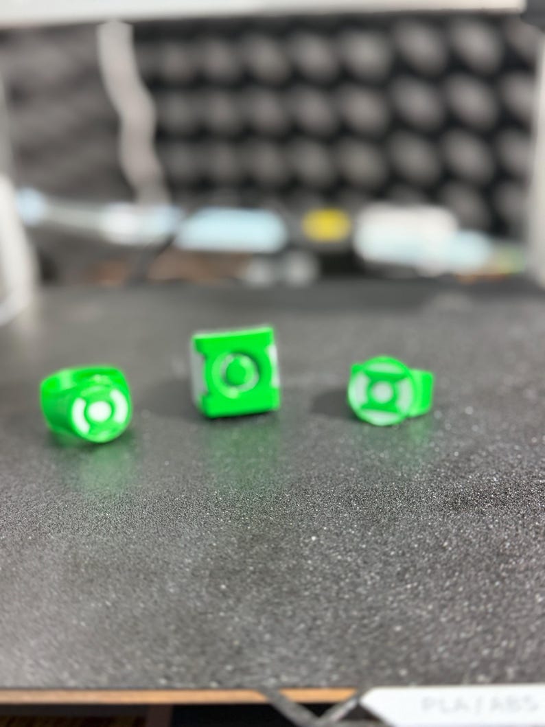 Puede incluir: Tres objetos impresos en 3D con el tema de Green Lantern, verdes y blancos. Los objetos incluyen dos anillos y un cubo, todos con el logotipo de Green Lantern. Los art&iacute;culos se muestran sobre una superficie gris texturizada.