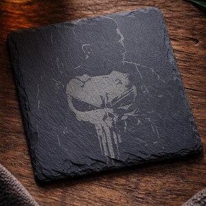 May include: Square black slate coaster with a gray graphic of a skull and the silhouette of a person. The coaster is on a wooden surface, with a glass of amber liquid in the background.