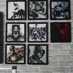 May include: A collection of eight square framed wall art prints featuring various comic book superheroes including Batman, Green Lantern, Wonder Woman, Superman, The Flash, and Green Arrow, arranged in a grid pattern on a light gray brick wall.