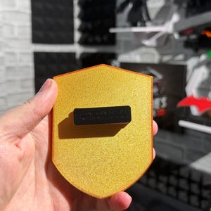 May include: A gold-colored shield-shaped magnet with a textured surface. The magnet has a black rectangular piece attached to the back with the text "CAUTION - MAGNETIC DEVICE DO NOT USE WITH PACEMAKER".