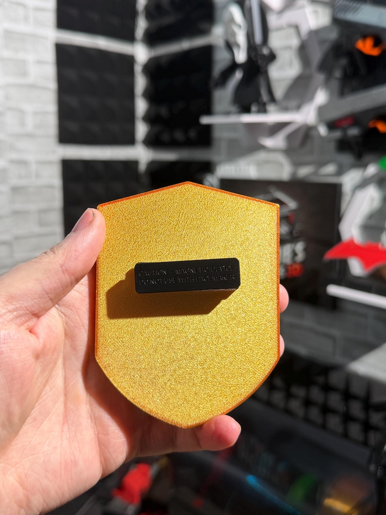 May include: A gold-colored shield-shaped magnet with a textured surface. The magnet has a black rectangular piece attached to the back with the text "CAUTION - MAGNETIC DEVICE DO NOT USE WITH PACEMAKER".