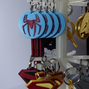 K&ouml;nnte beinhalten: Schl&uuml;sselanh&auml;nger mit Superhelden-Logos, darunter Spider-Man und Superman. Die Spider-Man-Schl&uuml;sselanh&auml;nger sind hellblau mit einem roten Spinnenemblem. Superman-Schl&uuml;sselanh&auml;nger sind rot und goldfarben. Die Schl&uuml;sselanh&auml;nger werden auf einem wei&szlig;en St&auml;nder pr&auml;sentiert.