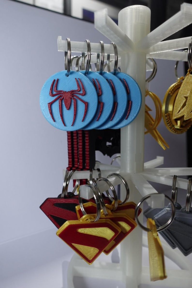 K&ouml;nnte beinhalten: Schl&uuml;sselanh&auml;nger mit Superhelden-Logos, darunter Spider-Man und Superman. Die Spider-Man-Schl&uuml;sselanh&auml;nger sind hellblau mit einem roten Spinnenemblem. Superman-Schl&uuml;sselanh&auml;nger sind rot und goldfarben. Die Schl&uuml;sselanh&auml;nger werden auf einem wei&szlig;en St&auml;nder pr&auml;sentiert.