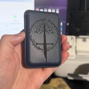 May include: A navy blue phone wallet featuring a tree and sword design. The design is surrounded by text. The wallet is rectangular and appears to be made of leather. The design is in black.