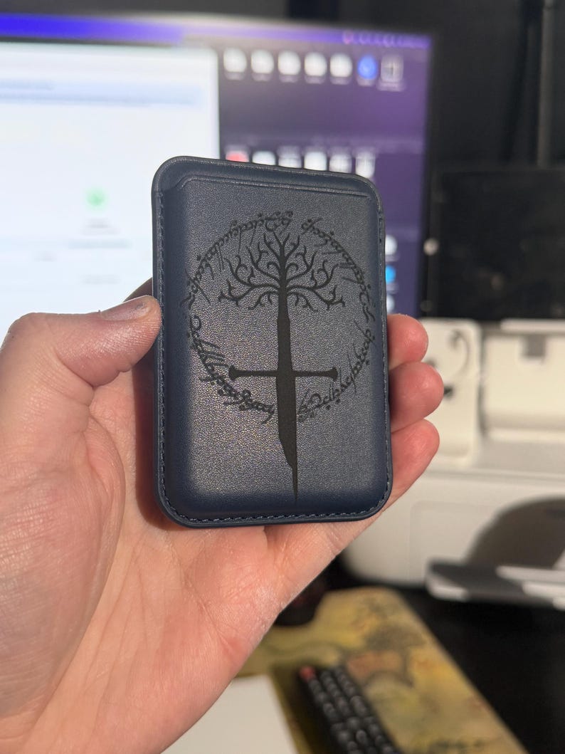 May include: A navy blue phone wallet featuring a tree and sword design. The design is surrounded by text. The wallet is rectangular and appears to be made of leather. The design is in black.
