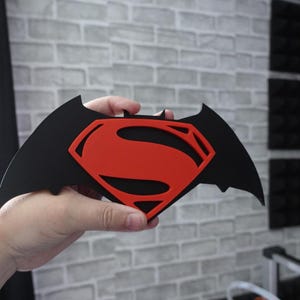 May include: A flat, black, bat-shaped emblem featuring a prominent red Superman logo cutout in the center. The design combines the iconic silhouette of a bat with the classic S-shield symbol, creating a layered, two-tone decorative piece.