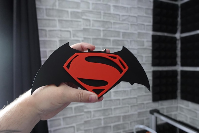 May include: A flat, black, bat-shaped emblem featuring a prominent red Superman logo cutout in the center. The design combines the iconic silhouette of a bat with the classic S-shield symbol, creating a layered, two-tone decorative piece.