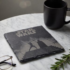 May include: A square, dark gray slate coaster with the Star Wars logo and a silhouette of a lightsaber battle. A black mug sits to the right. The coaster is on a white marble surface.