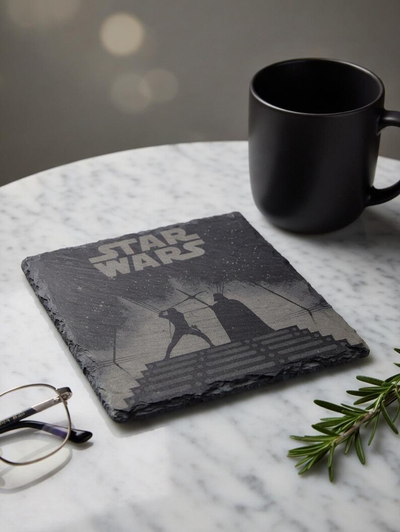 May include: A square, dark gray slate coaster with the Star Wars logo and a silhouette of a lightsaber battle. A black mug sits to the right. The coaster is on a white marble surface.