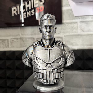 May include: A silver-colored bust of a man wearing a tactical vest with a skull emblem. The bust is 3D-printed and sits on a circular base. The background includes a sign that reads "Richie's Lens 3D".