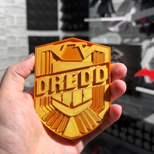 May include: A gold-colored, shield-shaped emblem with the word "DREDD" in bold, raised lettering. The emblem has a detailed, textured design with geometric patterns and is held in a hand.