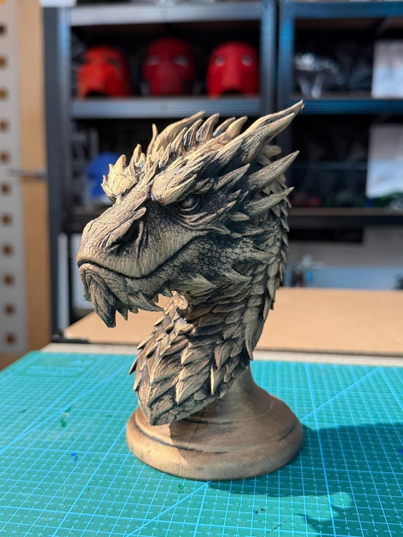 May include: A detailed, hand-painted dragon head sculpture in shades of brown and tan. The dragon has a textured, scaled appearance with sharp details, including pointed ears and a stern expression. The sculpture sits on a round base.